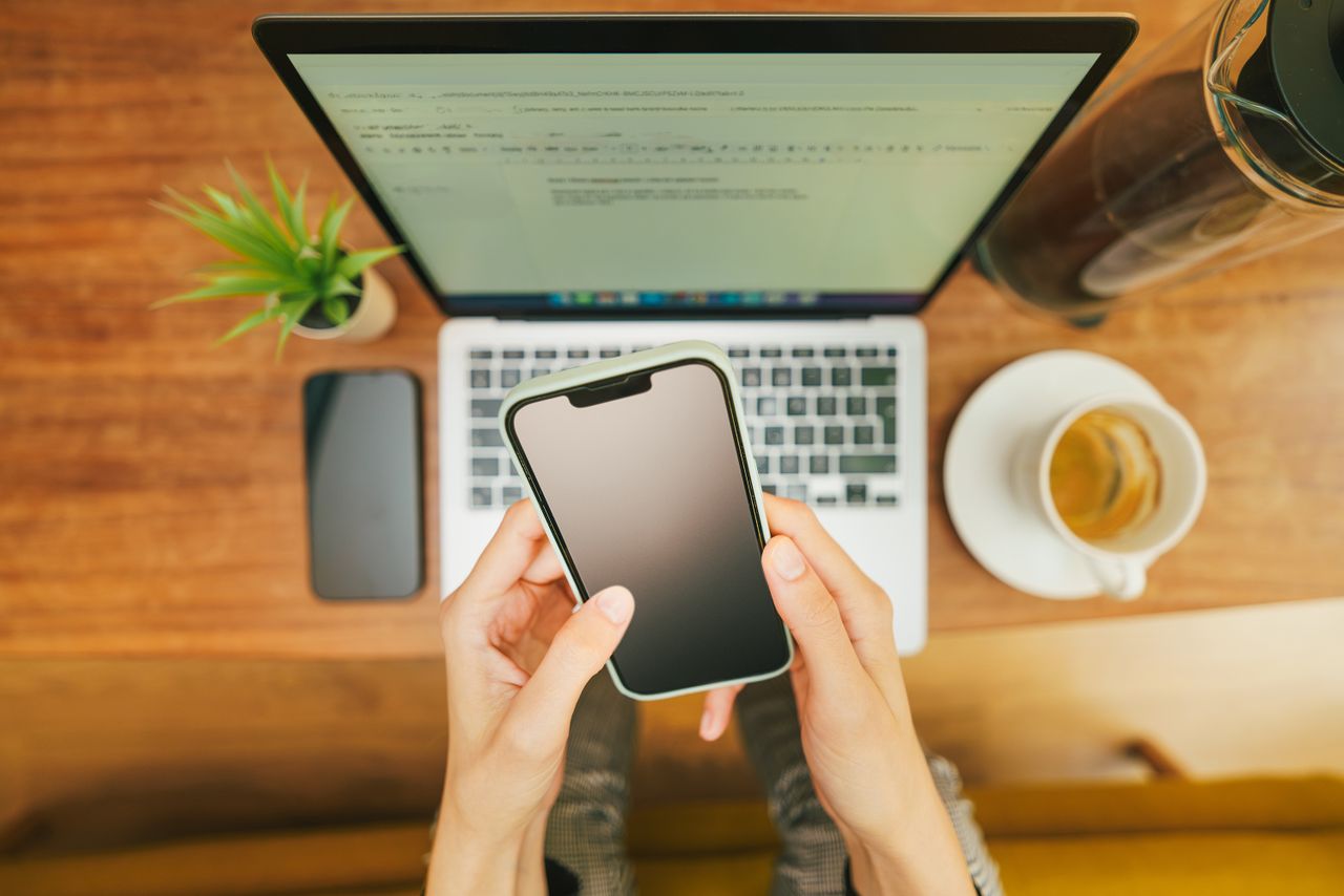 creative-approach-img Hands holding a smartphone over a laptop with coffee in a cozy home office setup.